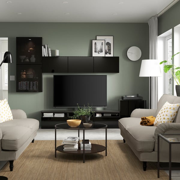 Современная гостиная со стенами цвета шалфея, тумбой под телевизор BESTÅ, серыми диванами, желтыми подушками, журнальным столиком с растением, торшером и телевизором с плоским экраном.