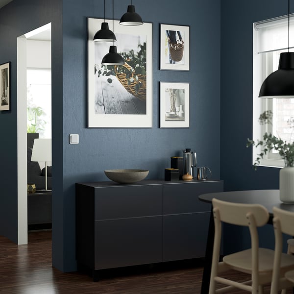 Modern dining room with dark blue walls, gray sideboard, white chairs, and black pendant lights. Gallery wall and window enhance minimalist design.