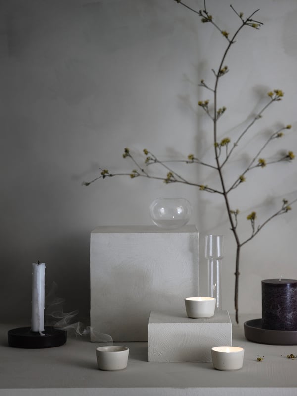 Minimalist setup: white ceramic holders, glass vase with branches, candles on stone plinth against grey wall.