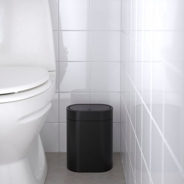 Black trash can in bathroom next to white toilet, designed to fit in small spaces.