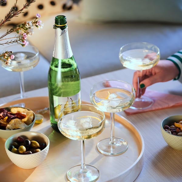 A group of elegant cocktails and appetizers on a table. Four glasses with clear drinks, a green bottle, and bowls of olives, cheeses, and nuts.
