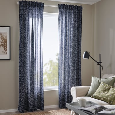 Dark blue floral curtains in room with light furniture, lamp, chair, cup & magazine.