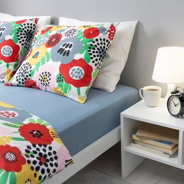 Bright bedroom: BRUKSVARA blue fitted sheet on bed, floral pillows, small white nightstand with coffee, books, alarm, lamp. Cozy, colorful decor.