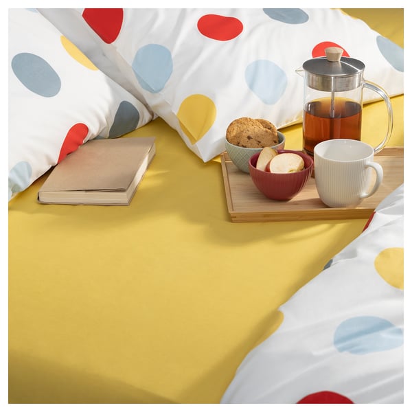 Cozy bedroom scene with BRUKSVARA yellow sheet, colorful polka dot pillow, book, coffee, and pastries.