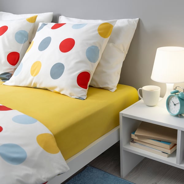 Bed with BRUKSVARA yellow sheet, polka dot pillows, white lamp, mug, books, and blue alarm clock.