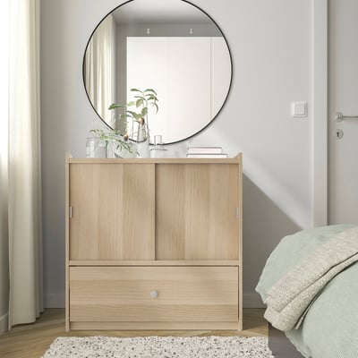 BRUKSVARA storage unit with light wood & sliding doors, smaller than bed & mirror, on rug near white bedspread.