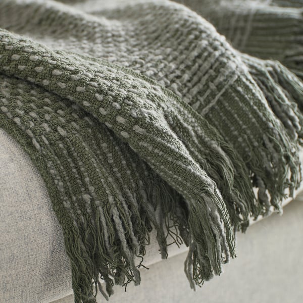 Soft handwoven blanket with fringes, textured dark grey-green pattern, displayed on a cushion.