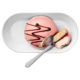 A pink dessert on a white plate with a spoon. Chocolate swirls top the treat.