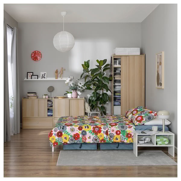 Bright, wooden bedroom with floral bedding and light gray walls. A wardrobe and a large plant.