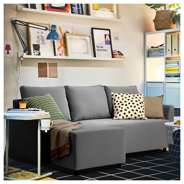 Modern grey l-shaped sofa with colorful cushions and throw, under shelf with books and lamp, black-tiled floor.