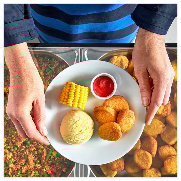 A person serves a plate with plant-based nuggets, corn, and ketchup over a counter.