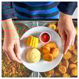 A person serves a plate with plant-based nuggets, corn, and ketchup over a counter.