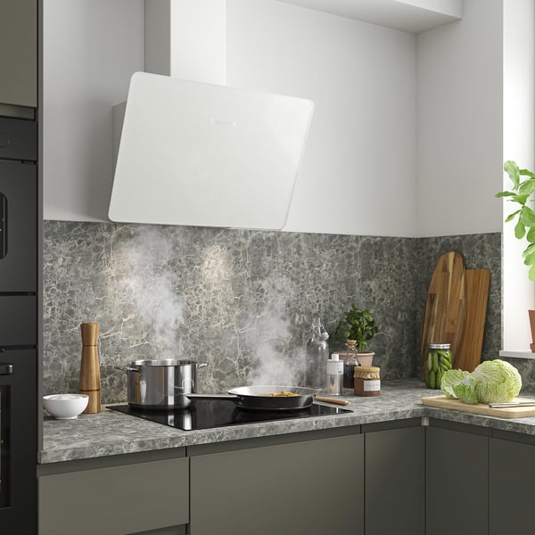 Modern kitchen with large white range hood, sleek marble counters, stove in use releasing steam, wooden utensils, and green vegetables.