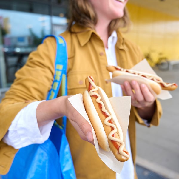 Osoba trzymająca dwa hot dogi z musztardą w papierze. Osoba ma na sobie musztardową kurtkę i niebieską torbę. Rozmyte tło.
