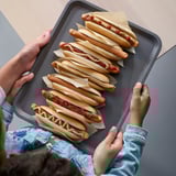 Dwie osoby, jedna osoba dorosła i jedno dziecko, trzymające tacę z hot dogami. Na tacy znajduje się sześć hot dogów pokrytych paskami ciasta, każdy zwieńczony keczupem.