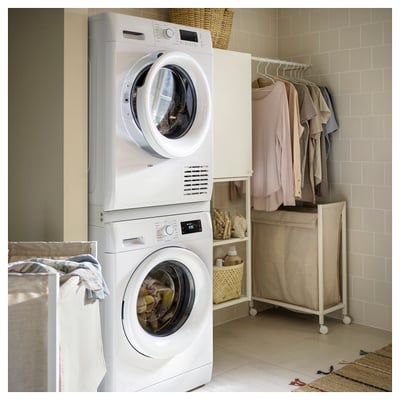 A white laundry room with stacked TYKARP appliances. Bottom: white washer, top: white dryer. Surrounding shelves hold clothes and laundry supplies.