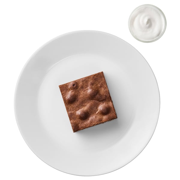 A square chocolate dessert on a white plate, with a small bowl of whipped cream beside it.