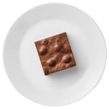 Square chocolate brownie on a white plate. Dense, rich texture with distinct chocolate flavor.