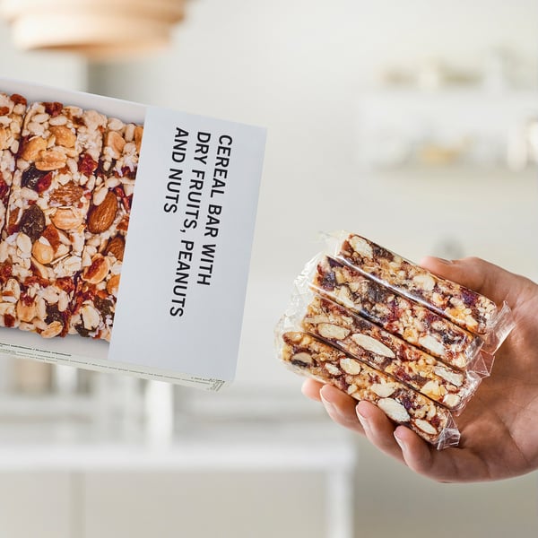 Hand holding packaged cereal bars, box nearby displaying product name and ingredients.