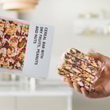 Hand holding packaged cereal bars, box nearby displaying product name and ingredients.