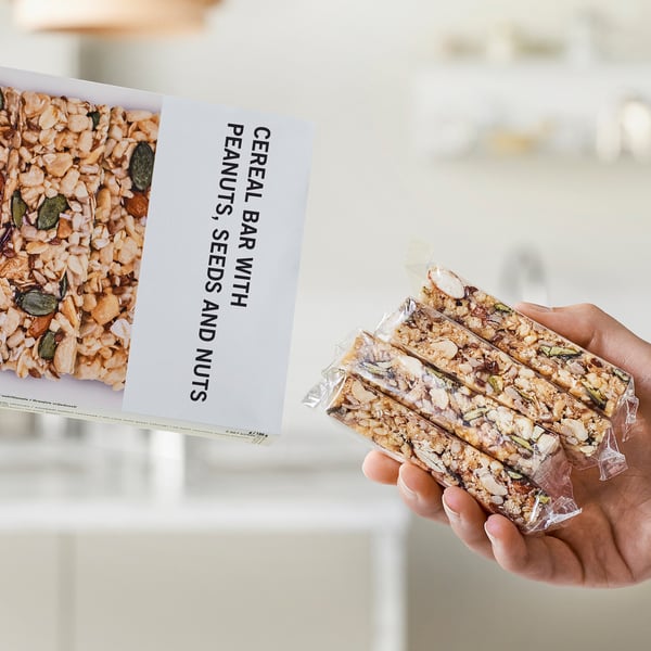 Hand holding three wrapped cereal bars next to box displaying image and text cereal bar with peanuts, seeds and nuts.