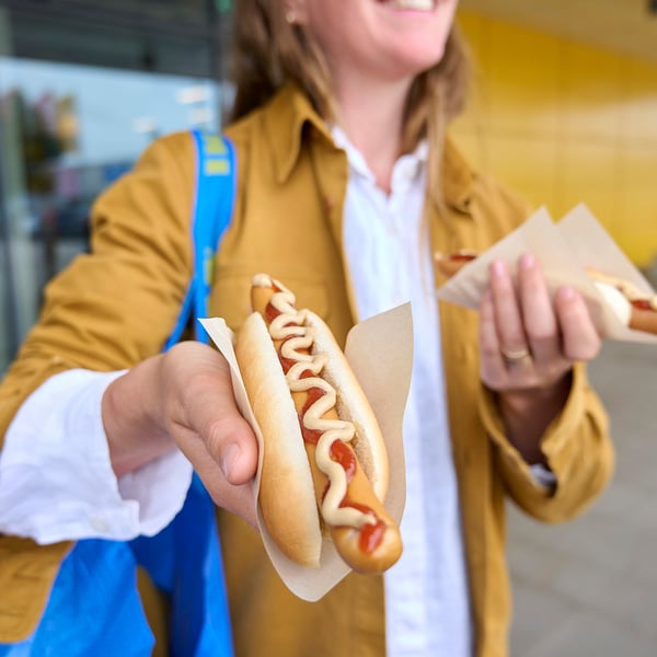 Person holding hot dog with mustard and ketchup in bread, wearing tan jacket with blue straps.