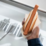A hand holds a hot dog wrapped in paper, showcasing the foods brown color and rounded shape, with a background of metallic surfaces.