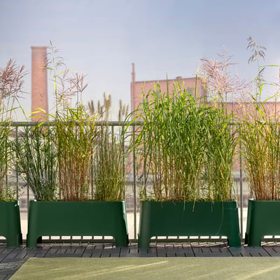 Three green aspern planters on a deck, filled with tall grasses, backed by red-brick buildings with smokestacks under a blue sky.