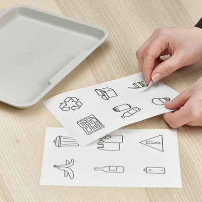 A person is applying clear stickers with waste sorting symbols onto a recycling bin. The stickers help identify what to throw in each bin.