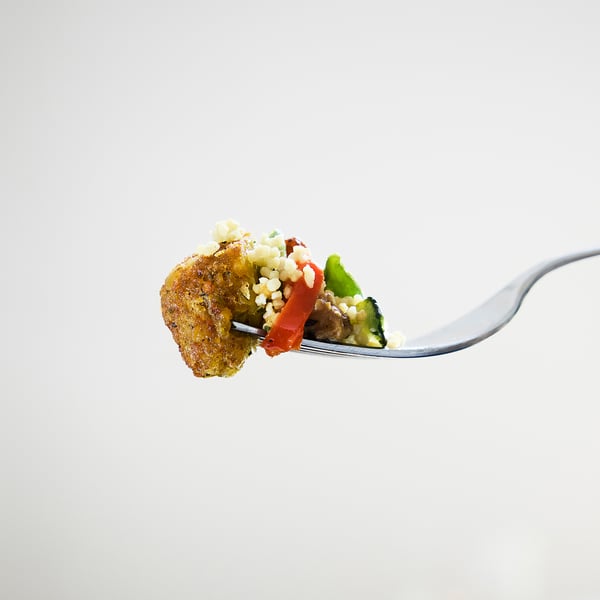 A fork holds a bite of falafel, couscous, and veggies against a white background.