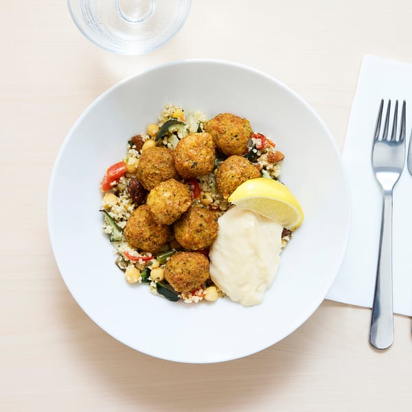 Falafel couscous bowl: golden falafels, couscous, veggies, aioli, lemon slice on white plate with fork.
