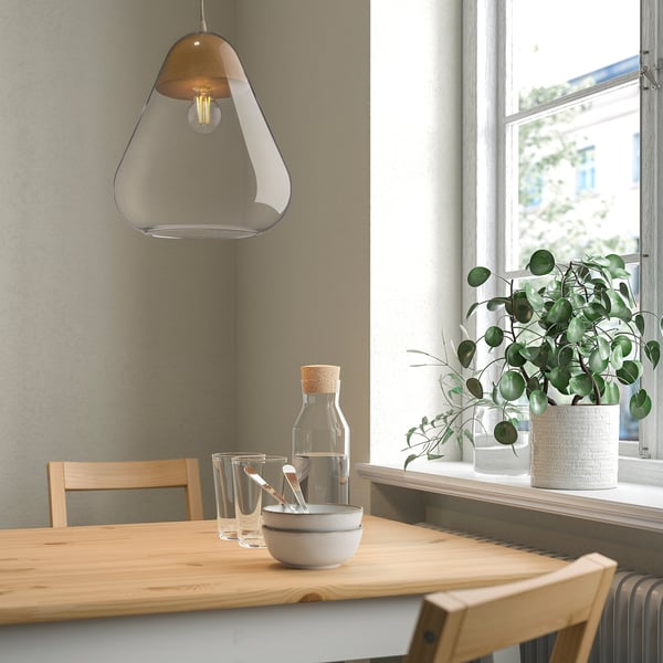 Lampa AKTERGAST ze szklaną kopułą nad drewnianym stołem, przy oknie z roślinami.