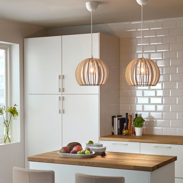 Lampy wiszące w kolorze beżowym wiszą nad wyspą kuchenną, kuliste z siatką, większe niż miska na owoce poniżej, białe szafki, wyłożony kafelkami backsplash.