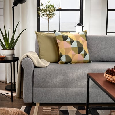 Przytulny salon z szarą sofą, kolorowymi poduszkami i lampą podłogową. Stolik boczny z roślinami i szyszkami. Ciepłe, naturalne światło.