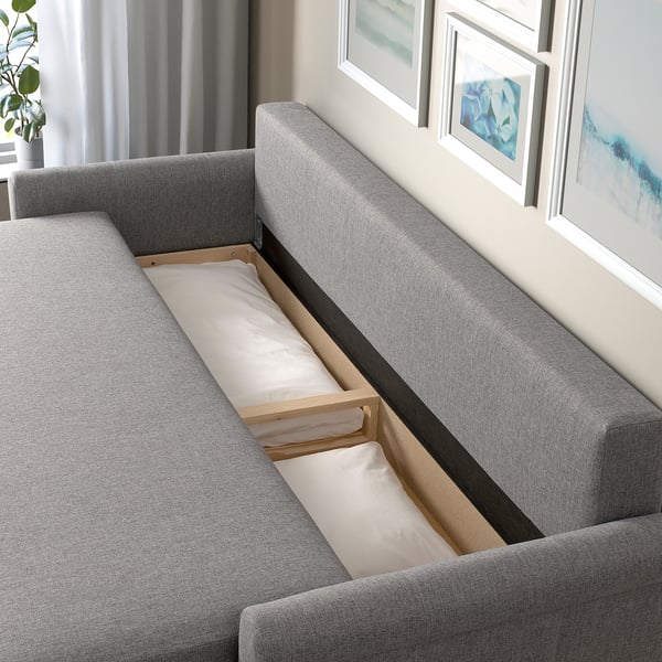 Szara rozkładana sofa DÅNHULT z otwartym schowkiem i poduszkami, umieszczona w pokoju obok sztuki i zasłon.