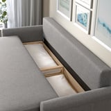 Szara rozkładana sofa DÅNHULT z otwartym schowkiem i poduszkami, umieszczona w pokoju obok sztuki i zasłon.
