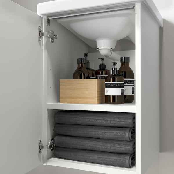 Otwórz białą szafkę łazienkową, w której na dolnej półce leżą starannie złożone szare ręczniki, a na górze przybory toaletowe.