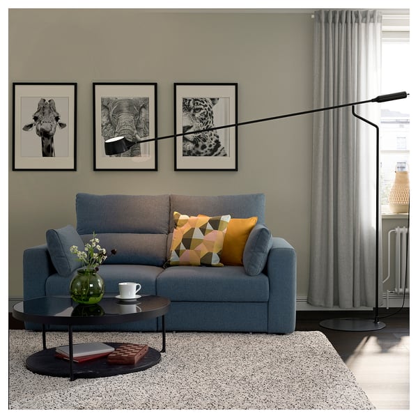 Nowoczesny salon z szarą sofą ebkilstuna, kolorowymi poduszkami, czarnym stolikiem kawowym, wydrukami na ścianie, zasłonami i lampą podłogową.