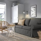 Nowoczesny salon z szarą sofą EVERTSBERG, krzesłem z jasnego drewna, białym stolikiem bocznym i szarym dywanem. Sofa jest duża i dominująca.