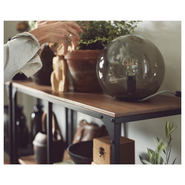 Osoba dostosowująca roślinę na stole FJÄLLBO z lampą i dekoracją.