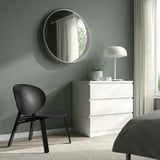 Minimalistyczna sypialnia z krzesłem, komodą, lustrem, lampą grzybkową, jasnozielonymi ścianami.