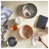 Kuchnia z narzędziami do pieczenia: waga, banneton, kubki, miód, łyżka, ściereczka, książka, skrobak do ciasta.