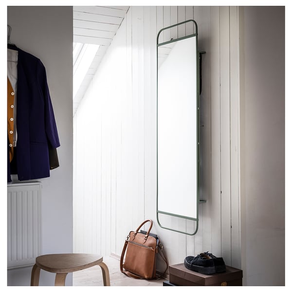 Zielone lustro GRANVÅG na białej ścianie z haczykami, stołkiem, torbą i ubraniami.