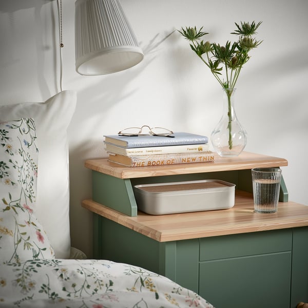 Stolik nocny z zielonym organizerem HARVMATTA, w którym znajdują się książki, lampka, kwiaty i szklanka.