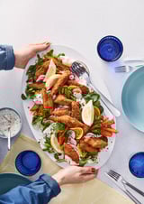 Półmisek chrupiących panierowanych kęsów ryb podawany z surówką. Ręce sięgające do służby. Niebieskie naczynia do picia i sztućce.