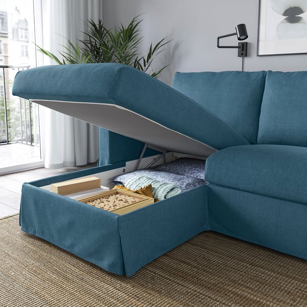 Niebieska sofa z podnoszonym szezlongiem, odsłaniającym schowek wypełniony kocami i pudełkiem.