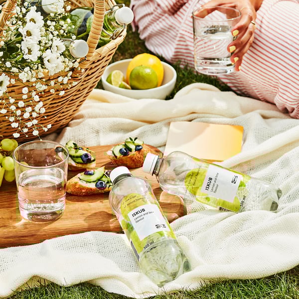 Scena piknikowa z butelką napoju cytrynowo-limonkowego ISKUB, szklankami wody, świeżymi owocami i przekąskami, rozłożonymi na kocu z kwiatami.