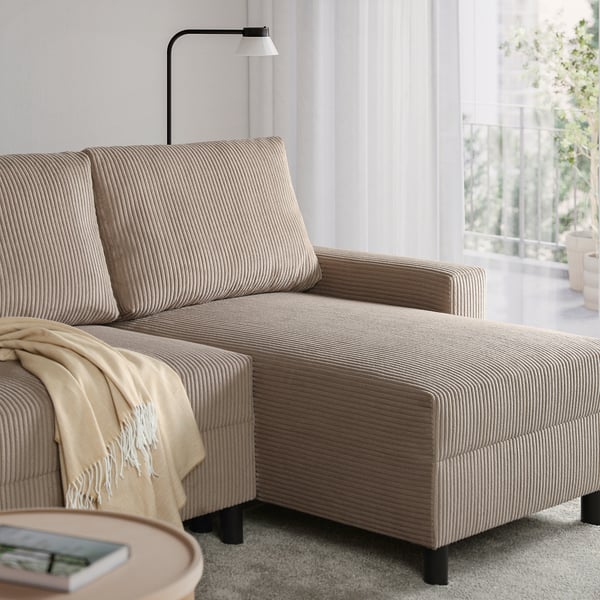 Beżowa sofa roczna z leżanką, poduszkami i kocem przy oknie. Okrągły stolik kawowy mieści książki i przerzuty, podkreślając nowoczesny wystrój.