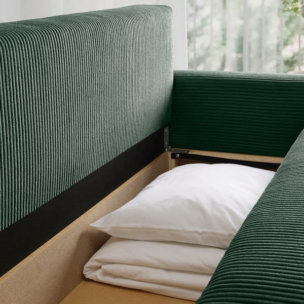 Zielona sofa z rozkładanym łóżkiem i schowkiem, białe poduszki.