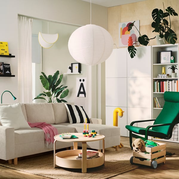 Salon z beżową kolumną JÄRINGSHOLM, drewnianym stolikiem kawowym, zielonym fotelem wypoczynkowym, żywym wystrojem, dużą rośliną, półką na książki i lampą wiszącą.